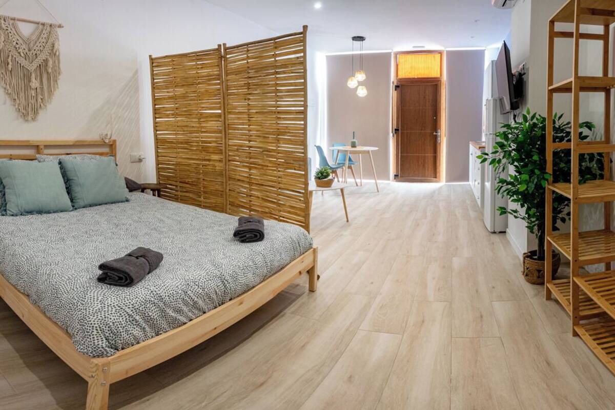 A bedroom with a wooden bed, a small stool, a macrame wall hanging, a bamboo room divider, and a wooden shelf unit.