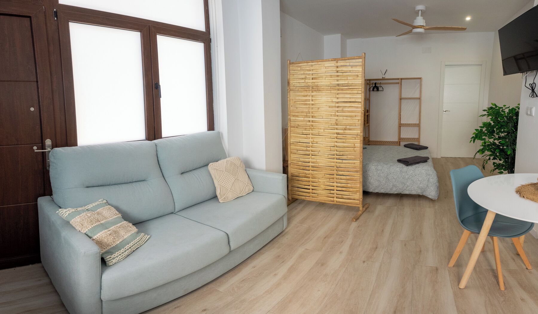 Una sala de estar moderna con un sofá azul claro, un separador de madera, una zona de comedor con una mesa redonda y un ventilador de techo.