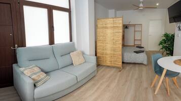 Una sala de estar moderna con un sofá azul claro, un separador de madera, una zona de comedor con una mesa redonda y un ventilador de techo.