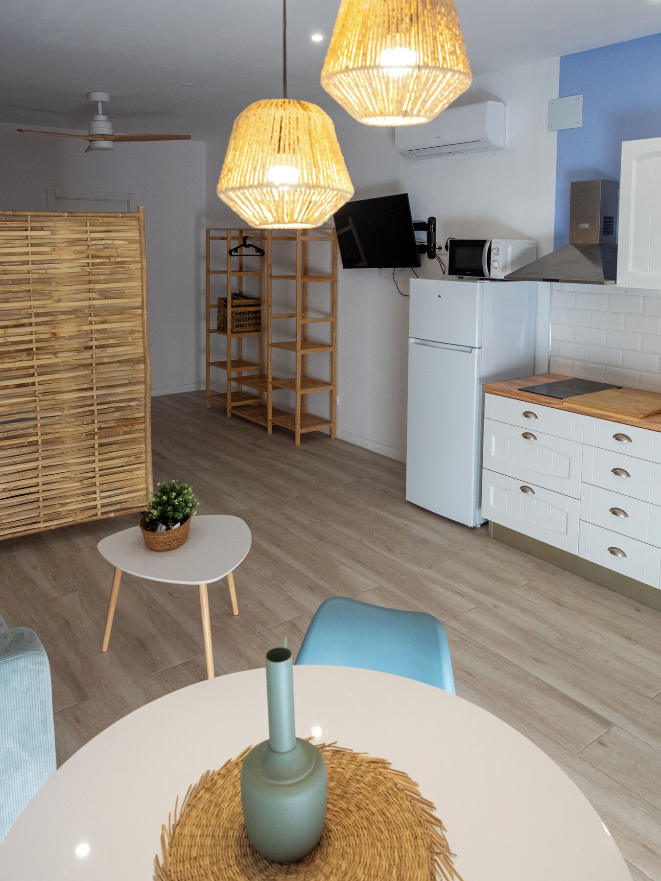 A modern kitchen with white cabinetry, a microwave, and a refrigerator. There are two hanging pendant lights, a small round table with a vase, and a blue chair.