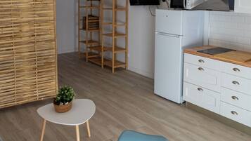 A modern kitchen with white cabinetry, a microwave, and a refrigerator. There are two hanging pendant lights, a small round table with a vase, and a blue chair.