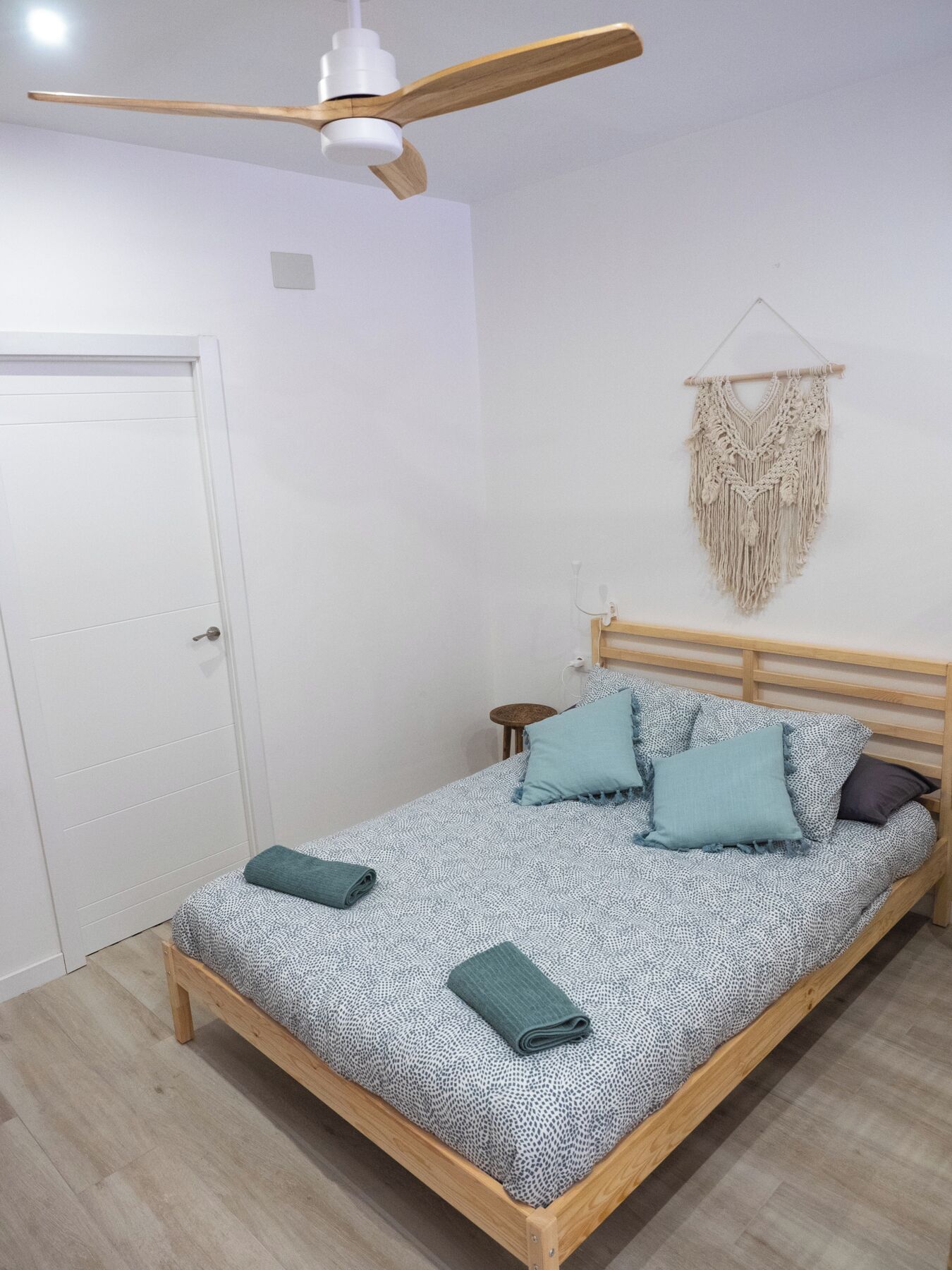 Habitació individual amb llit de fusta, ventilador al sostre, penjoll macramè a la paret i porta blanca.