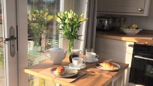 A breakfast setup with a teapot, cups, a bowl of fruit, and pastries on a wooden table by a window with a view of greenery. - STUNNING SHEPHERDS HUT WITH HOT TUB IN BEAUTIFUL RURAL CHESHIRE, NEAR NANTWICH (Nantwich)