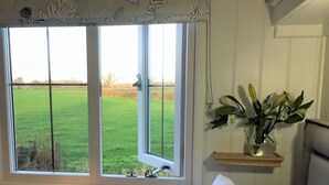 A room with a large window offering a view of a green field. There is a wooden shelf with a vase of flowers and a small table. - STUNNING SHEPHERDS HUT WITH HOT TUB IN BEAUTIFUL RURAL CHESHIRE, NEAR NANTWICH (Nantwich)