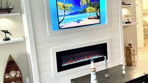 A modern living room with a flat-screen TV mounted above a fireplace, a wooden shelf with decorative items, and a dark coffee table. - Beach Front Condo with Breathtaking Views (Jensen Beach)