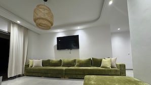 A modern living room with a green sofa, a flat-screen TV mounted on the wall, and a woven light fixture. - Family apartment with private garden, swimming pool and close to the beach - Cabo Negro (Cabo Negro)