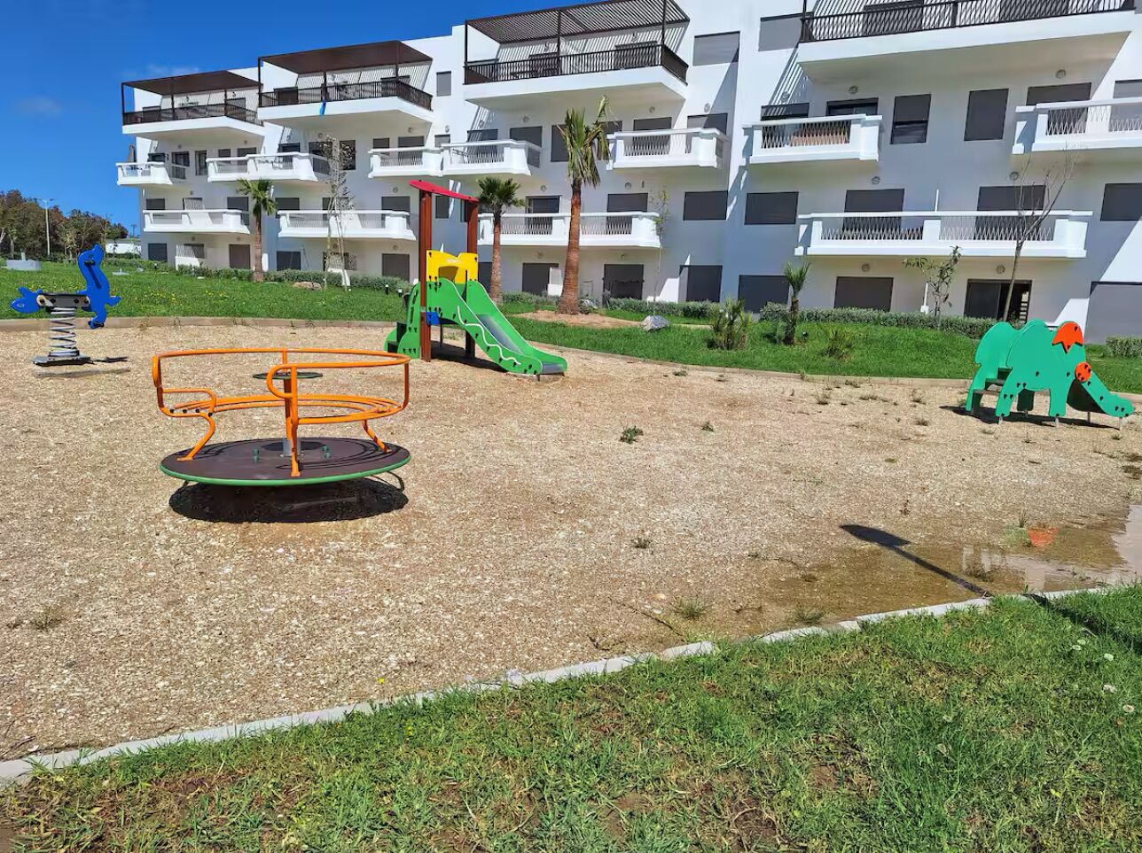 A playground with a seesaw, slide, and climbing structure, surrounded by a building with balconies and a grassy area.