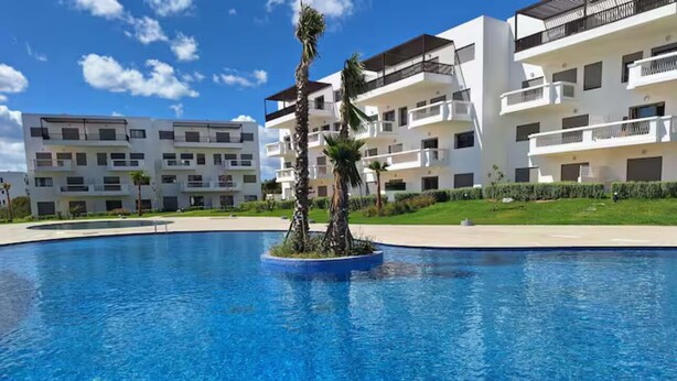 A swimming pool with a small island in the center, surrounded by modern buildings with balconies. - Family apartment with private garden, swimming pool and close to the beach - Cabo Negro (Cabo Negro)