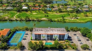 Vista aérea de una zona residencial con un edificio grande que cuenta con piscina y cancha de tenis, rodeado de vegetación y un lago.