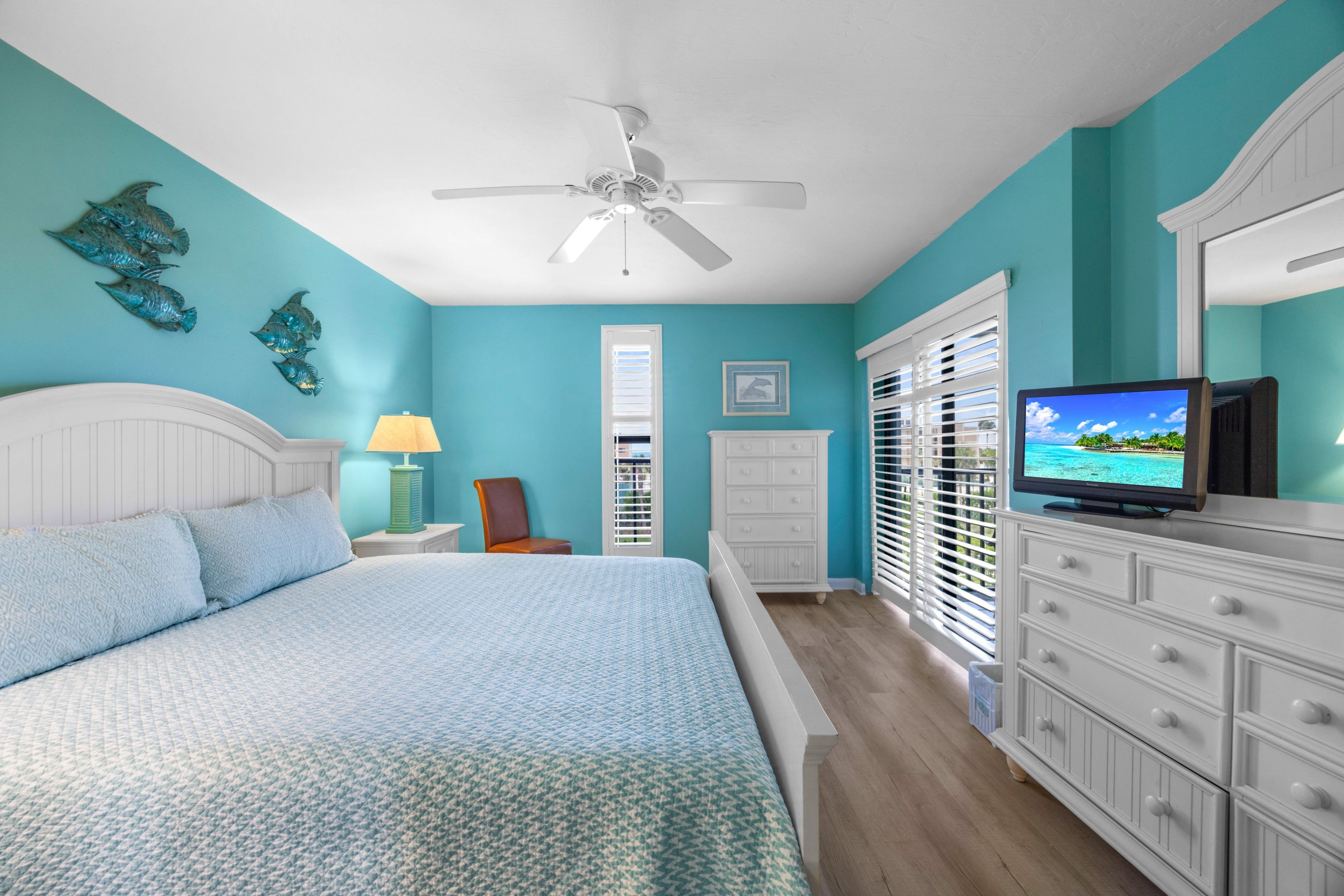 Une chambre à coucher avec un lit, un téléviseur, un ventilateur de plafond et une fenêtre avec des stores.