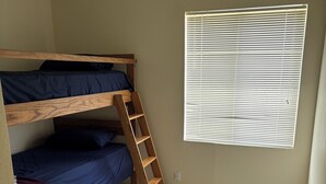 A room with a bunk bed, a wooden ladder, a window with blinds, and a wooden floor.
