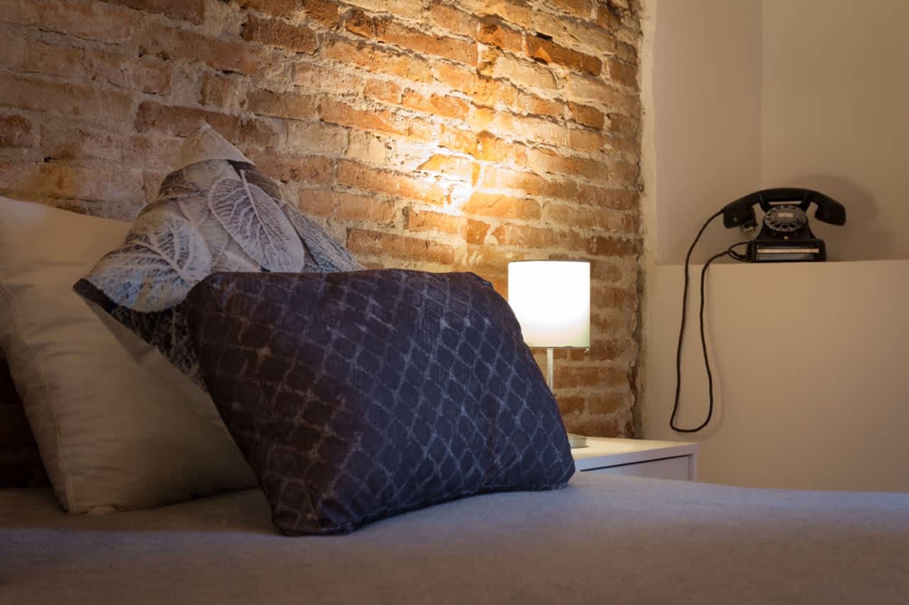 A bedroom with a brick wall, a bed with patterned pillows, a bedside lamp, and a vintage telephone.