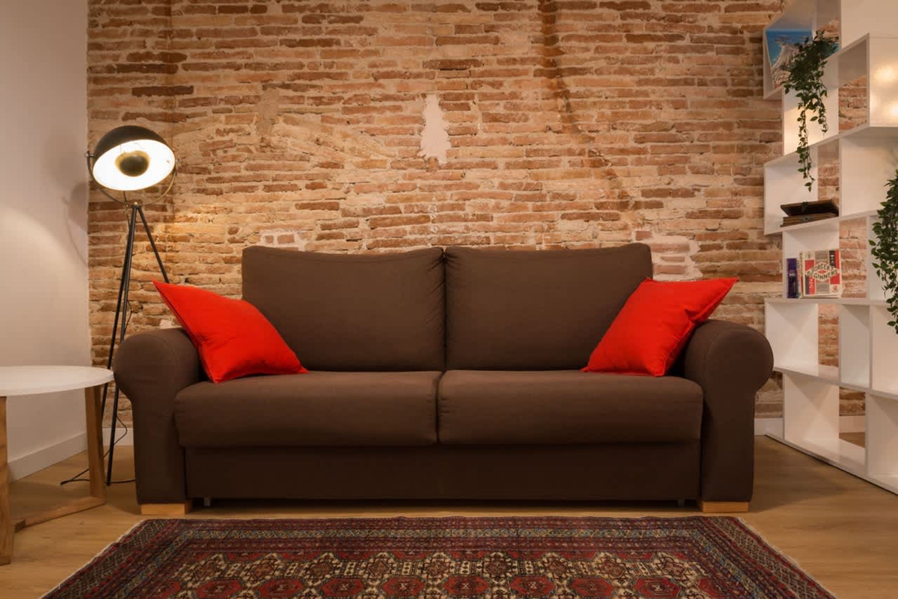 Un sofá marrón con cojines rojos, una mesa redonda, una lámpara de pie y una alfombra estampada en una habitación con pared de ladrillo y estanterías blancas.