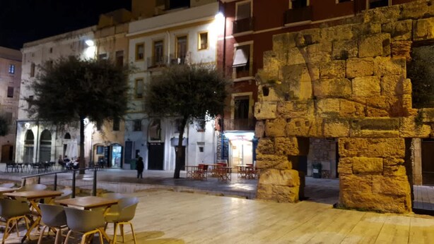 Un área de café al aire libre con mesas y sillas, un muro de piedra y edificios al fondo.