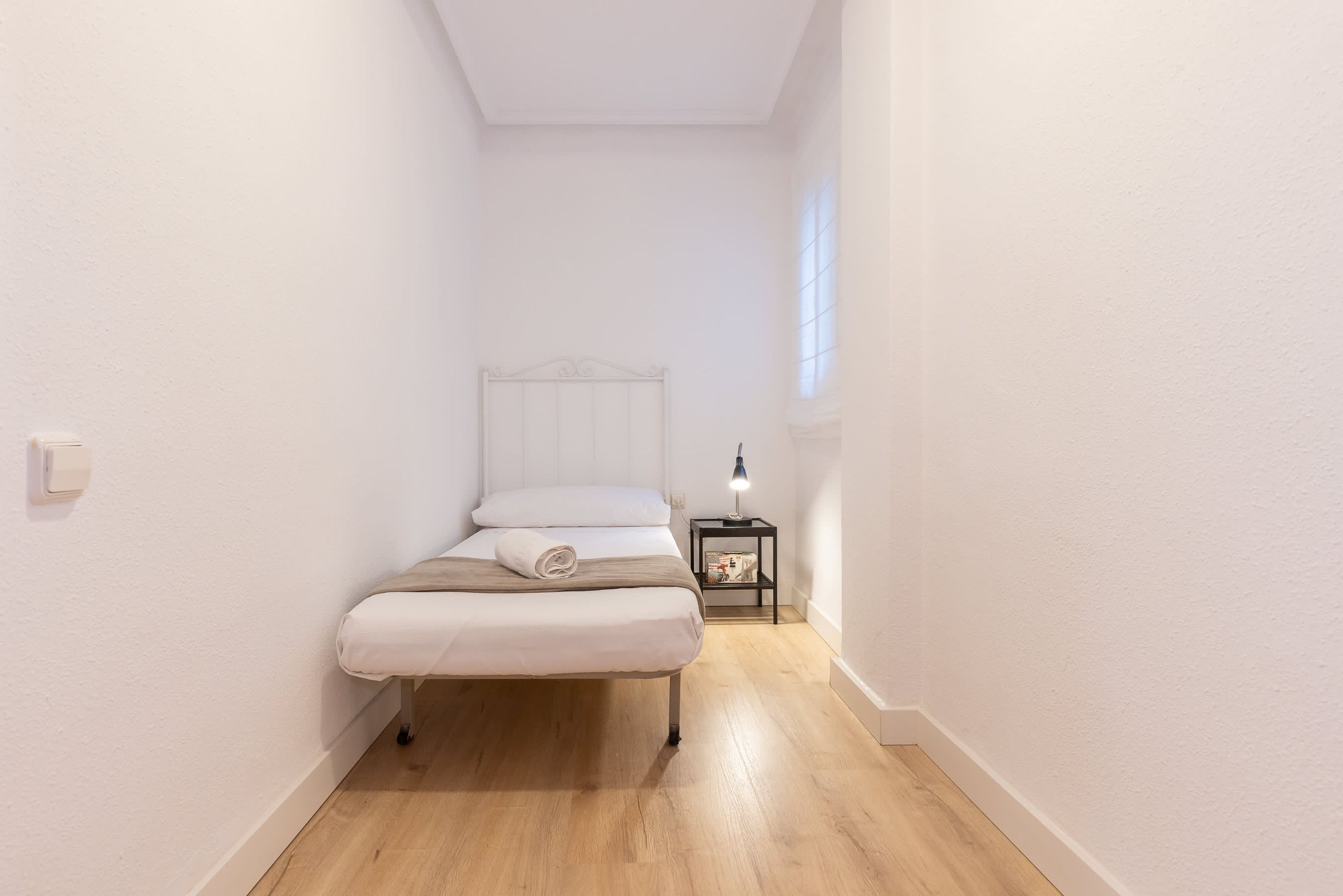 Una habitación estrecha con una cama, una mesita de noche y una pequeña mesa.
