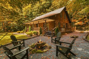 Eine Blockhütte mit Veranda, umgeben von einer gepflegten Schotterterrasse und einem Feuerkorb, eingebettet in einen bewaldeten Bereich.