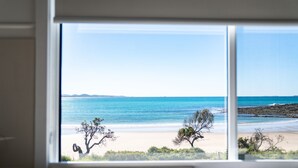 Vista a la playa a través de una ventana, con mar tranquilo, orilla arenosa y vegetación escasa.