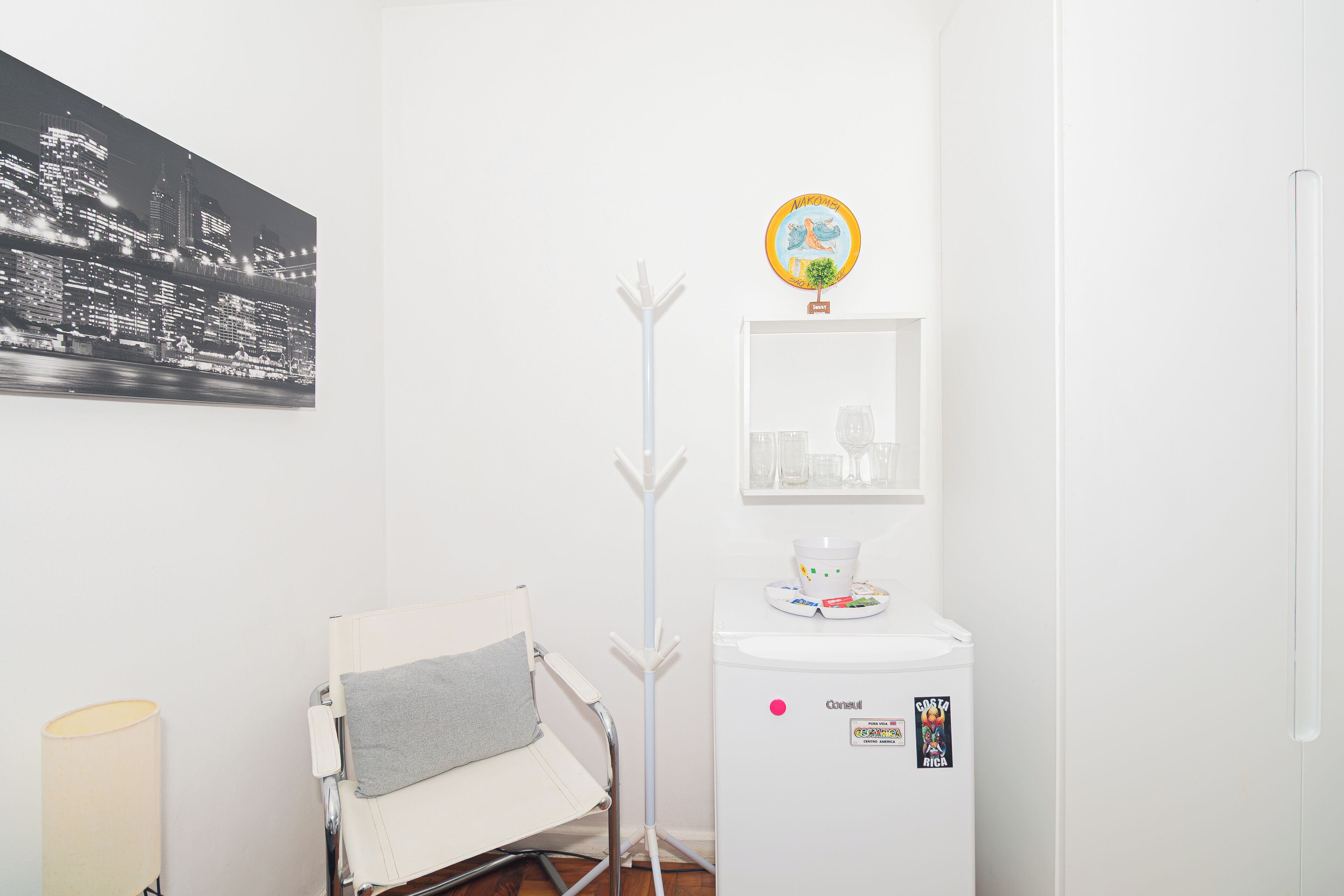 Een hotelkamer met een witte stoel, een staande lamp, een koelkast en een ingelijst schilderij aan de muur.