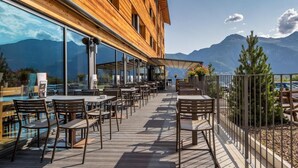 En udendørs terrasse med træborde og stole, store vinduer og udsigt til bjerge.