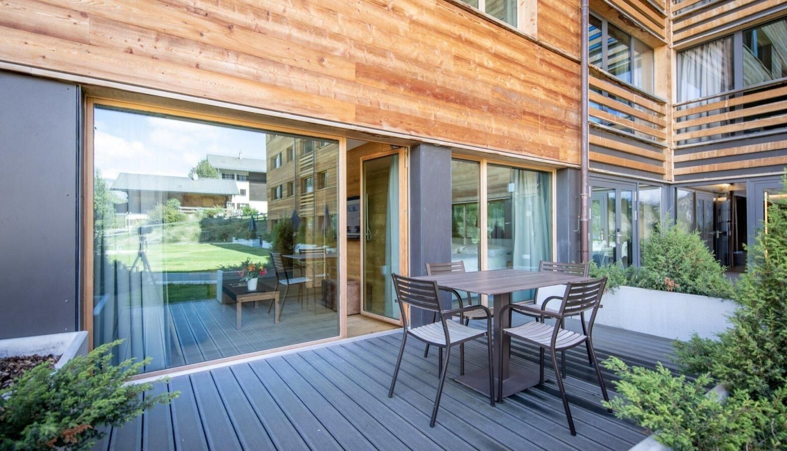 A modern outdoor patio with a wooden deck, a glass door leading to a garden, and a table with chairs.
