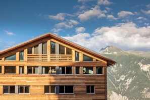 A wooden chalet with a triangular roof, large windows, and a mountain backdrop.