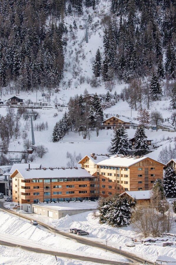A large wooden building with multiple floors, likely a hotel or resort, surrounded by snow-covered trees and a ski lift in the background.
