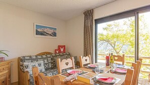 A dining area with a wooden table set for four, a sofa with patterned cushions, a small wooden cabinet, and a window with a view of trees and mountains.