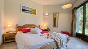 A bedroom with a bed, bedside lamps, a nightstand, a window with curtains, and a wall-mounted mirror.