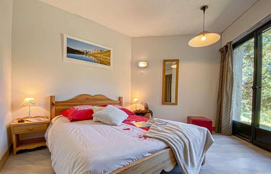 A bedroom with a bed, bedside lamps, a nightstand, a window with curtains, and a wall-mounted mirror.