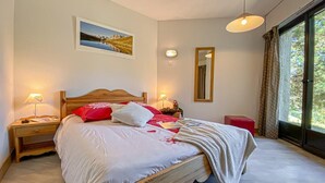 A bedroom with a bed, bedside lamps, a nightstand, a window with curtains, and a wall-mounted mirror.