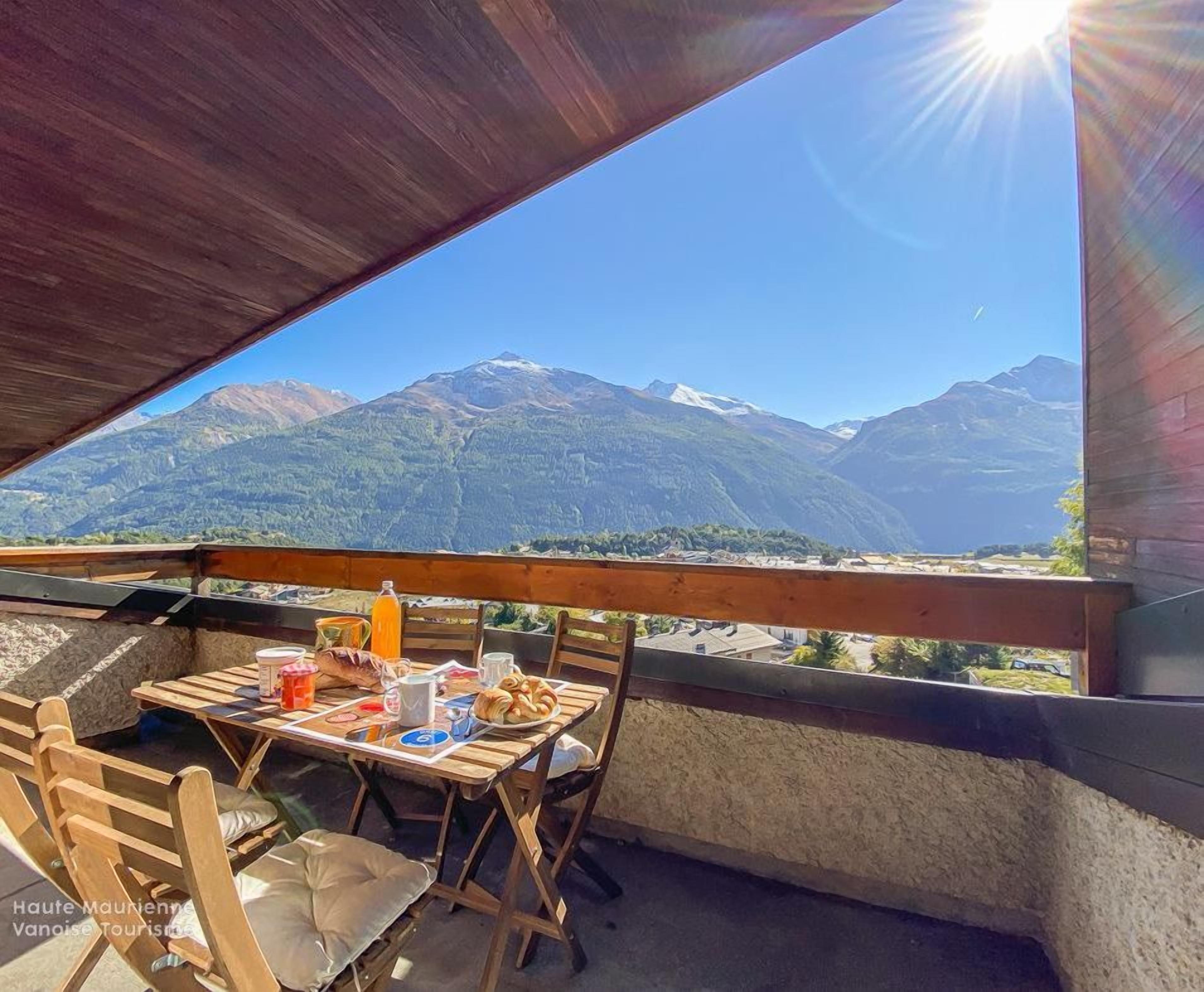 Une table en bois, entourée de chaises, sur un balcon donnant sur un paysage montagneux.