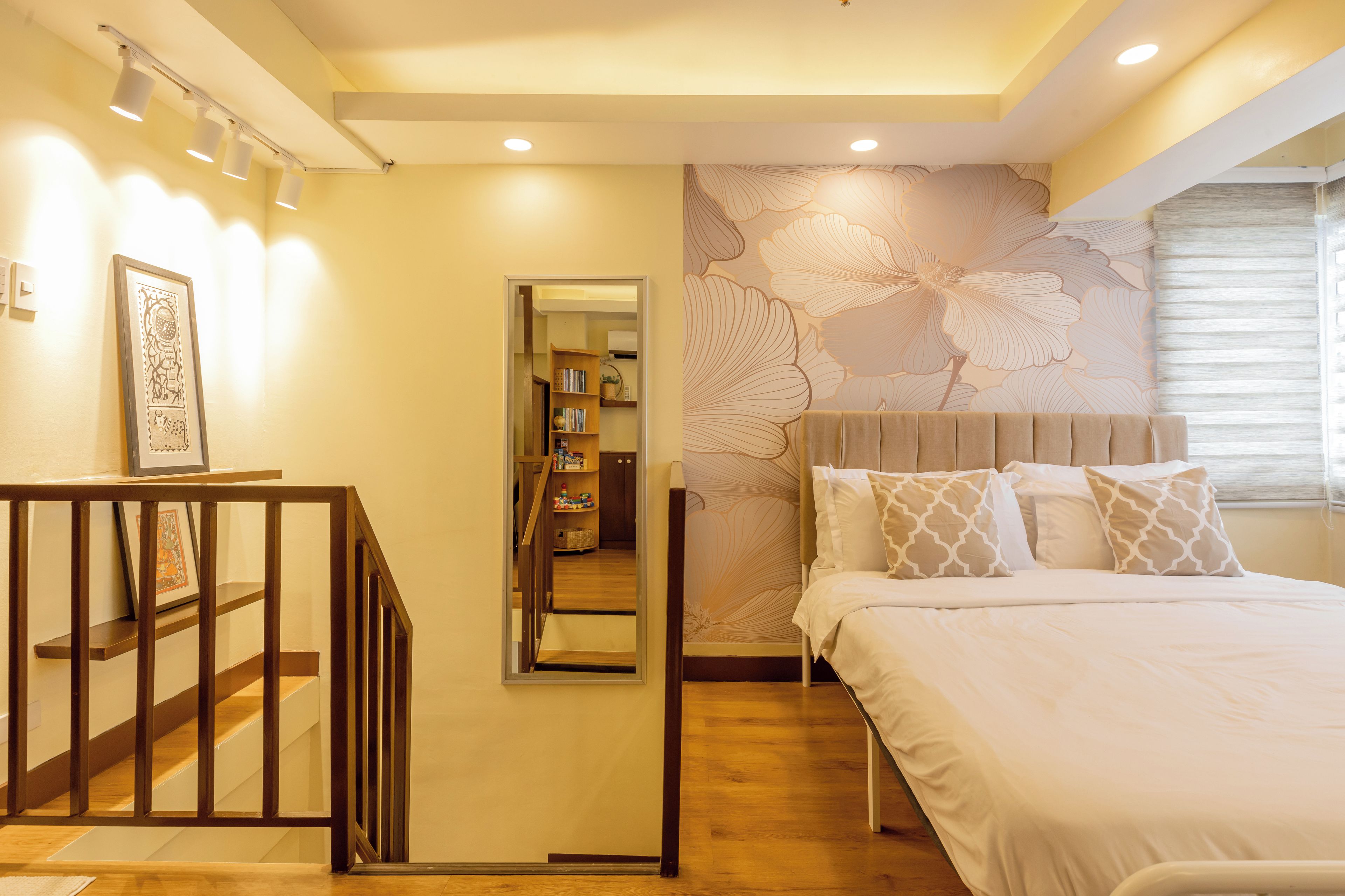 A bedroom with a bed, a wooden staircase, a mirror, and a wall with a floral pattern.