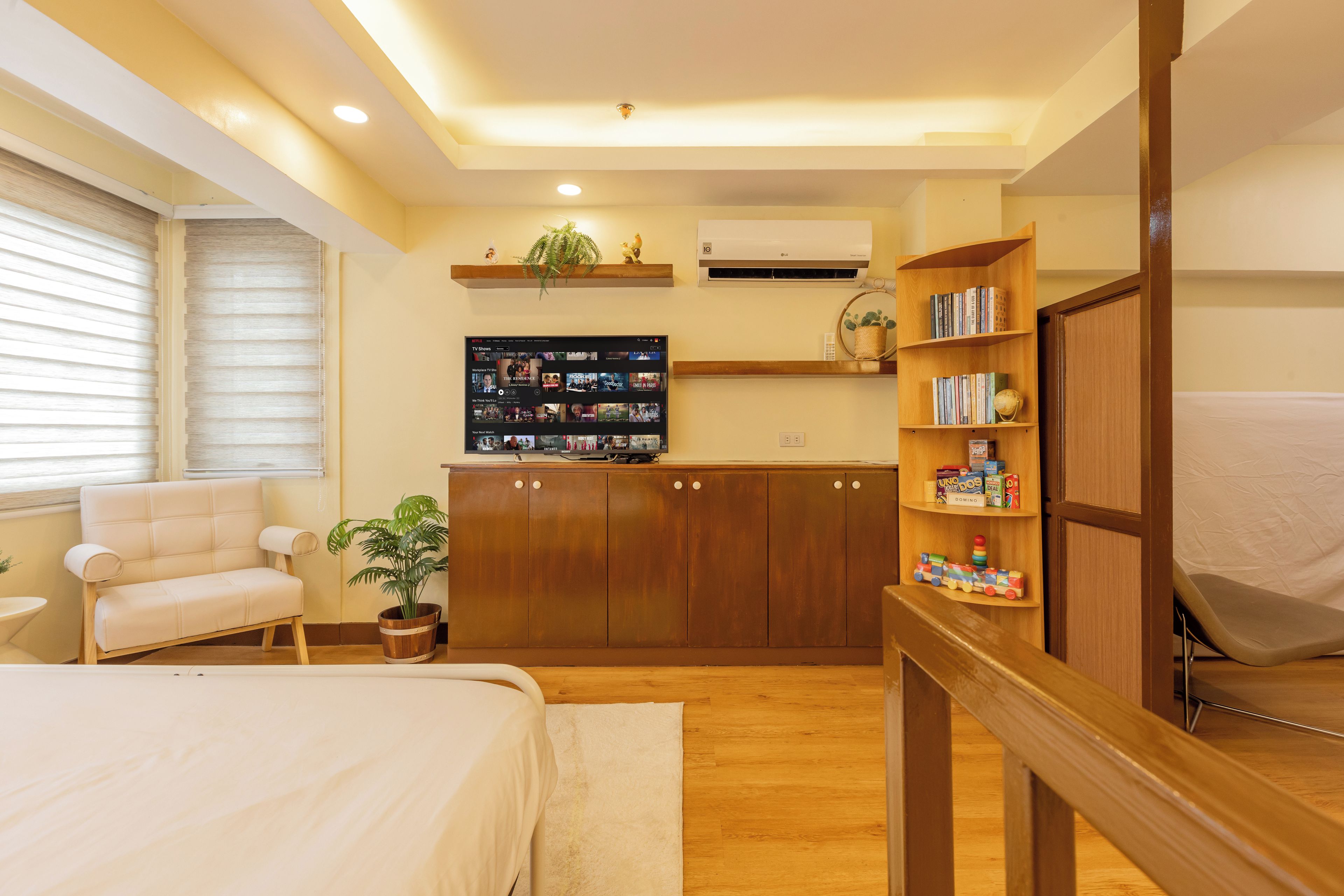 Un dormitorio con una cama, una silla, una planta en maceta, un televisor montado en la pared y un armario de madera.