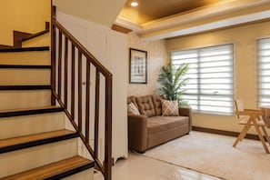 Una escalera con barandillas de madera que conduce a una sala de estar con un sofá, una planta en maceta y una ventana con persianas.