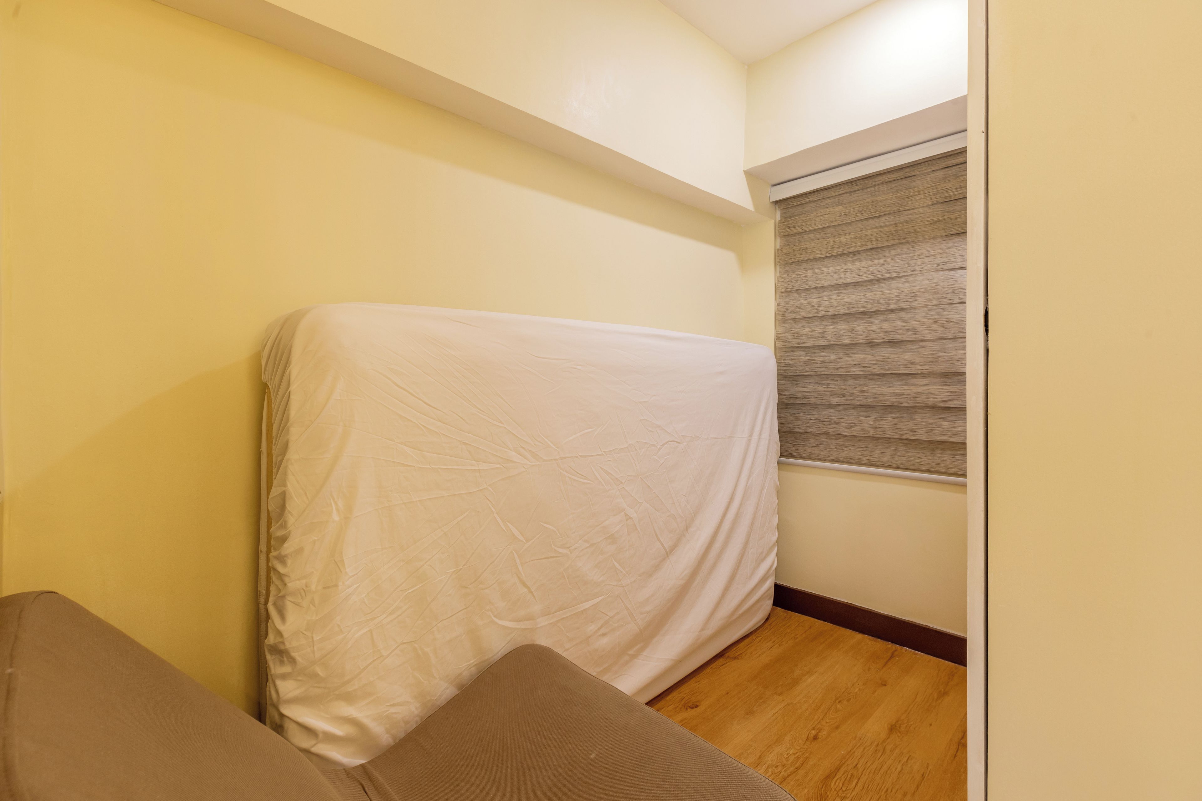 Una habitación con un colchón cubierto por una sábana blanca, piso de madera y una ventana con persianas.
