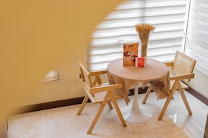 Una mesa redonda pequeña con mantel tejido, dos sillas de madera, un atril para menús y un florero con plantas secas.