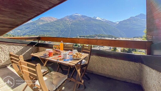 Une table en bois, entourée de chaises, sur un balcon donnant sur un paysage montagneux.