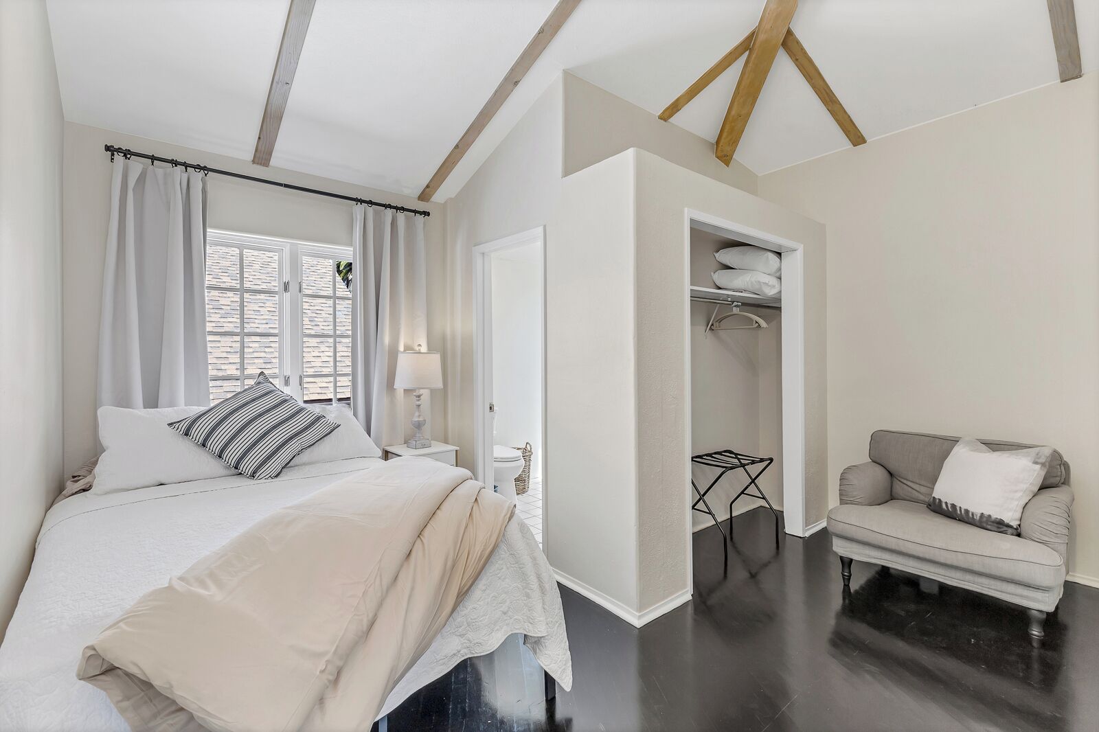 A bedroom with a bed, a nightstand, a lamp, a window with curtains, a closet, and a chair.