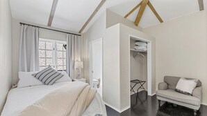 A bedroom with a bed, a nightstand, a lamp, a window with curtains, a closet, and a chair.