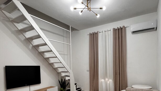 A modern living room with a herringbone wood floor, a sofa, a TV on a white console, a staircase, and a ceiling light fixture. - Cozy and well-maintained house in the historic center of Catania (Catania)