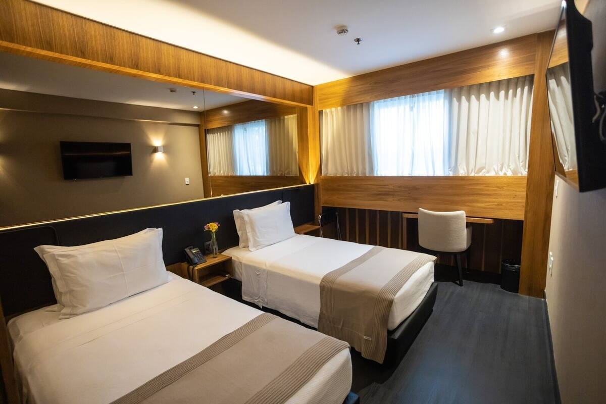 A hotel room with two beds, a desk, a chair, a TV, and a window with curtains.