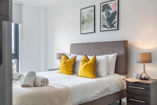 A neatly made bed with white and yellow pillows, a bedside table with a lamp, and two framed artworks on the wall.