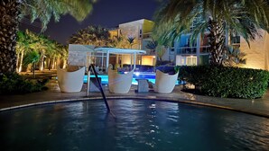 A nighttime view of a resort with a swimming pool, palm trees, and a modern building with a covered seating area.