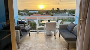 A balcony with wicker furniture, a table, and a sofa, overlooking a residential area with houses and a clear sky at sunset.
