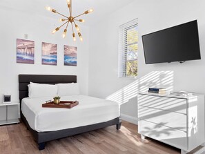 A modern hotel room with a bed, bedside table, TV, artwork, and a unique ceiling light fixture.