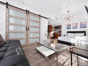 A modern hotel room with a sliding glass door closet, a glass coffee table with books, a bed with white linens, and a kitchenette in the background.