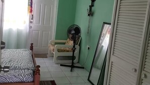 Ein Zimmer mit einem Bett, einem Sessel, einem Standventilator, einer Tür und einem an der Wand montierten Telefon.