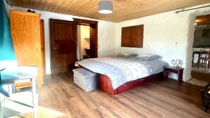 Ein geräumiges Zimmer mit Holzbalkendecke, einem Bett mit gemustertem Überwurf, einem kleinen Tisch und einem Sessel.