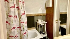 Un baño con una bañera, una mesita blanca con artículos de aseo, y una cortina de ducha con diseños de flamencos.