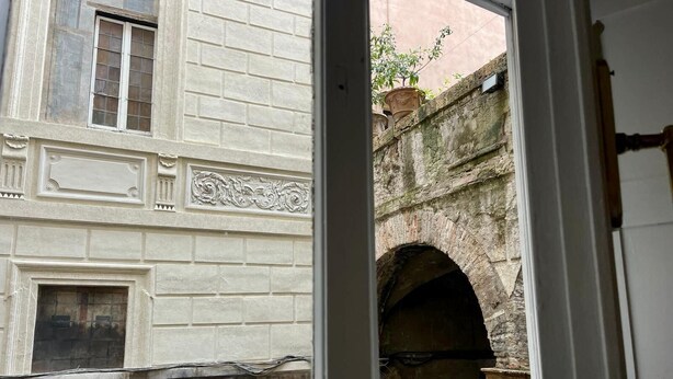 Vista a través de un vano de puerta que muestra un edificio con un arco de piedra y un ventanal con marco decorativo.